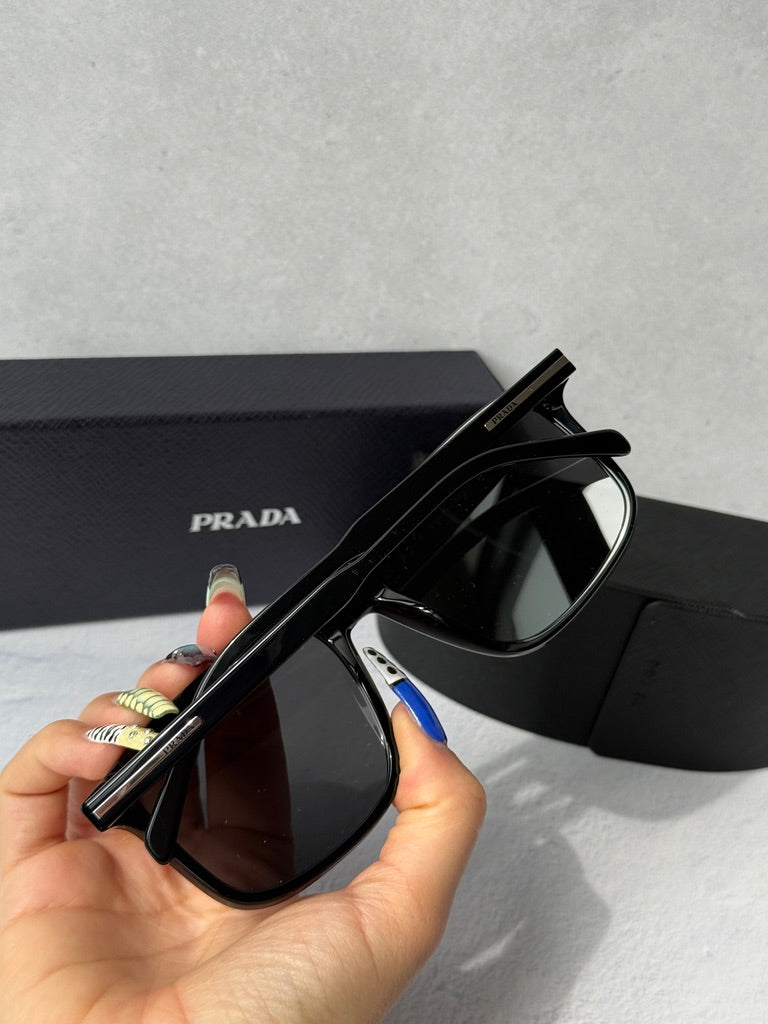 Black aviator sunglasses with dark lenses, PRADA temple logo visible, held in hand
