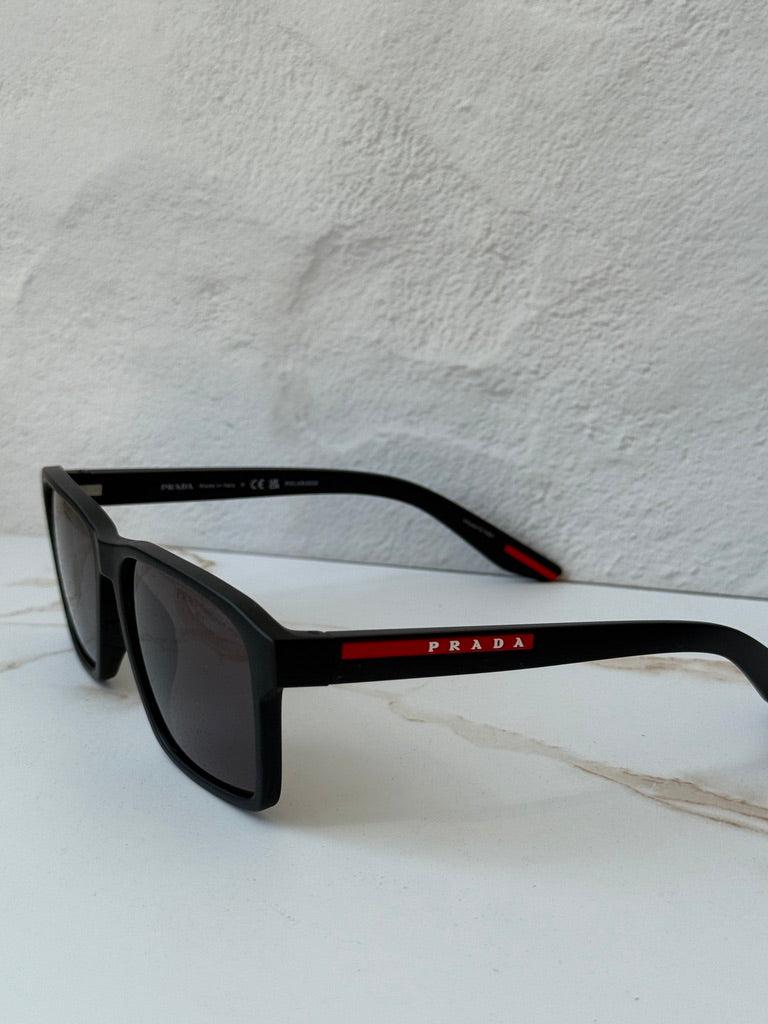 Black polarized sunglasses, angled view; dark lenses and red Prada logo bar on arm