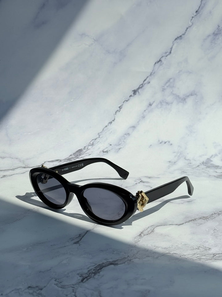 Black oval sunglasses with dark lenses and gold logo hinge, angled view