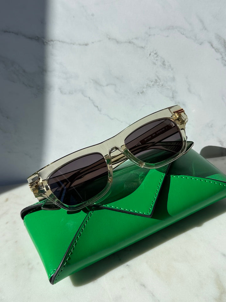 Mitre rectangular sunglasses with clear frame, gold accents, dark lenses, on green case