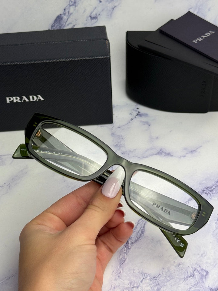 Transparent green eyeglass frames held by hand, rectangular lenses, Prada logo on temple.