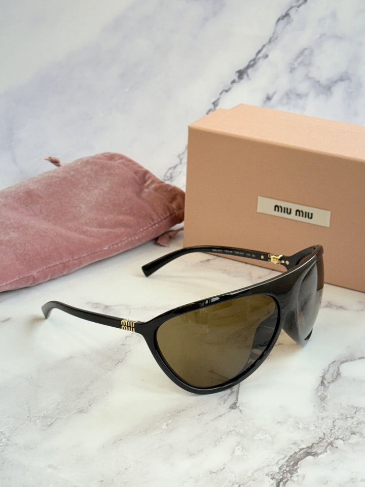 Black shield sunglasses with brown lenses near a pink cloth pouch and beige box.