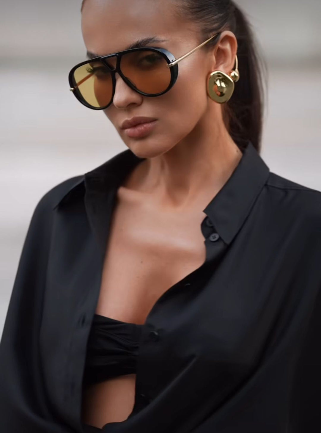 Black aviator sunglasses with gold drop earrings on model, yellow-tinted lenses.