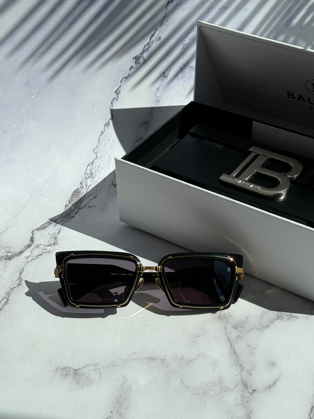 Balmain black square sunglasses with gold frame beside branded case