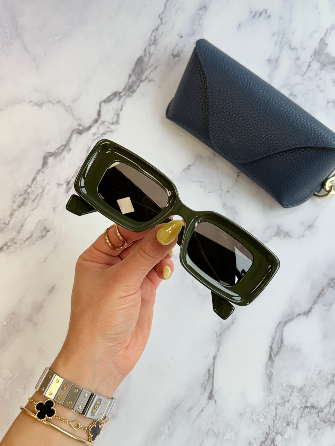 Dark green square sunglasses in hand with blue zip case beside them