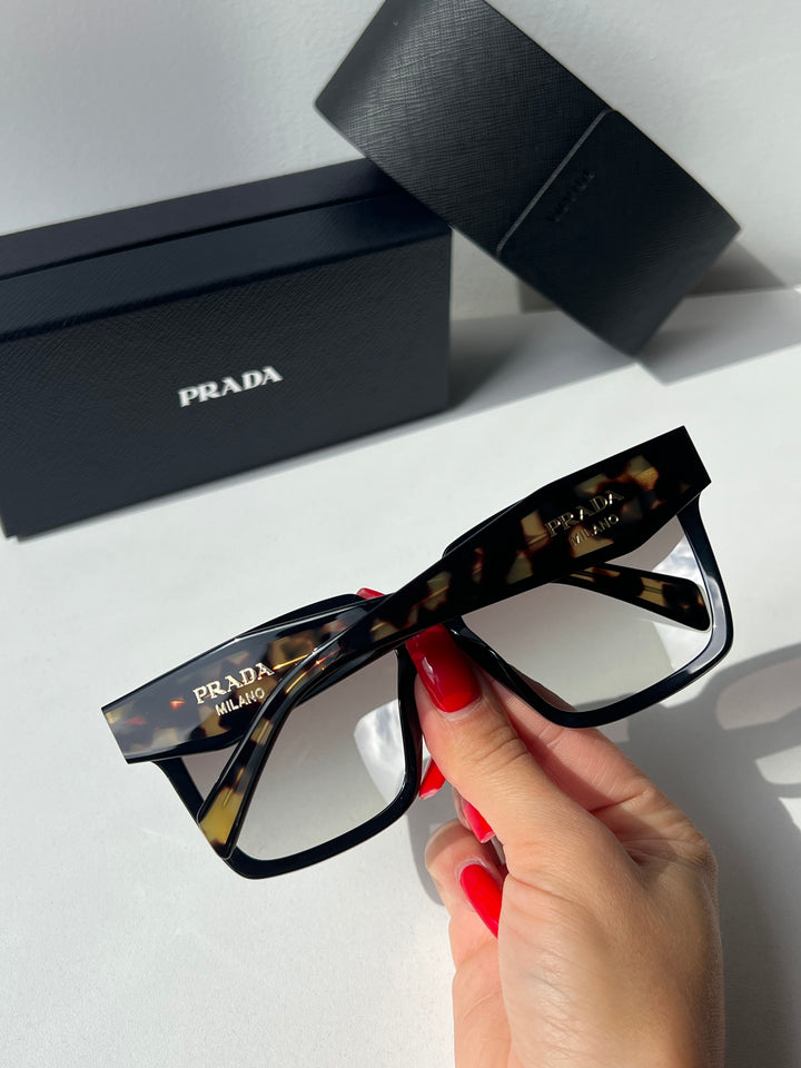 Prada oversized sunglasses held by hand, black havana frame with gold temple text