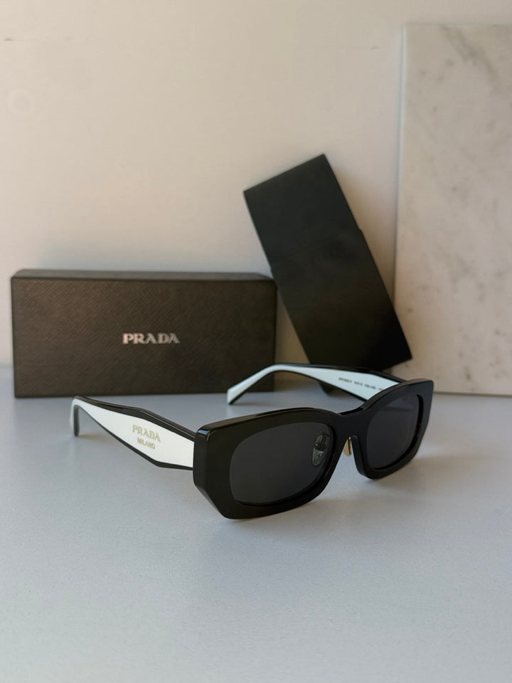Prada black sunglasses with dark lenses, white temple arms and case packaging.