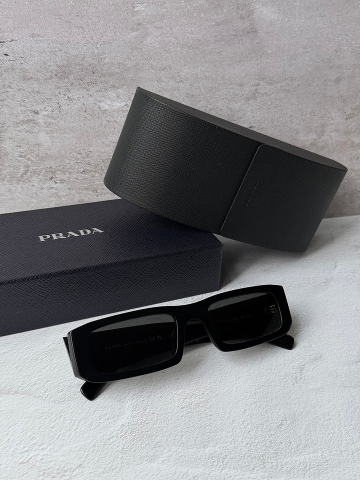 Prada black sunglasses with dark lenses, shown beside a matching case and box.
