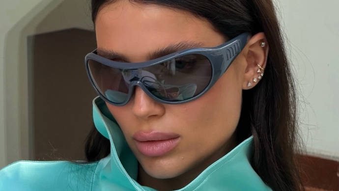 A close-up of a model wearing oversized dark gray shield sunglasses and a shiny light aqua high-collar zip-up jacket.