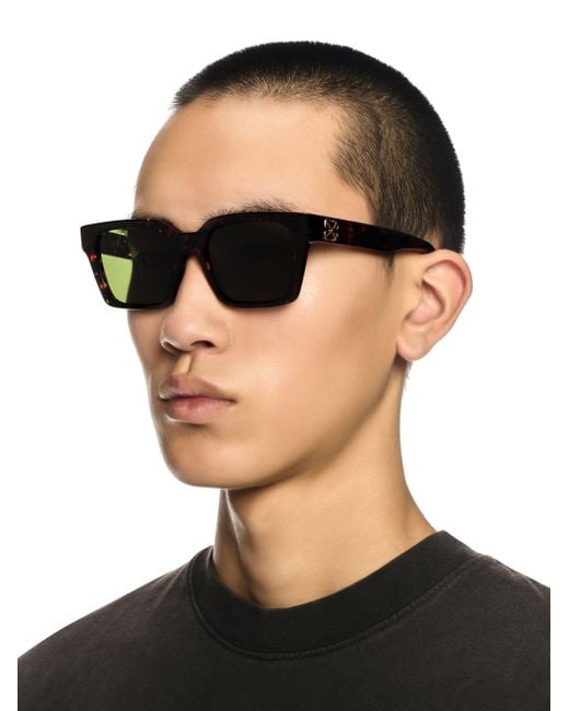 Black square sunglasses with dark lenses worn by model, logo on side arms